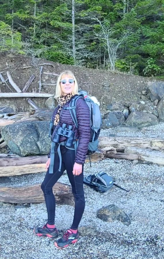 Maria Faires, RD and Boat Captain, standing on a scenic beach wearing a technical hiking backpack, representing active travel and outdoor exploration.